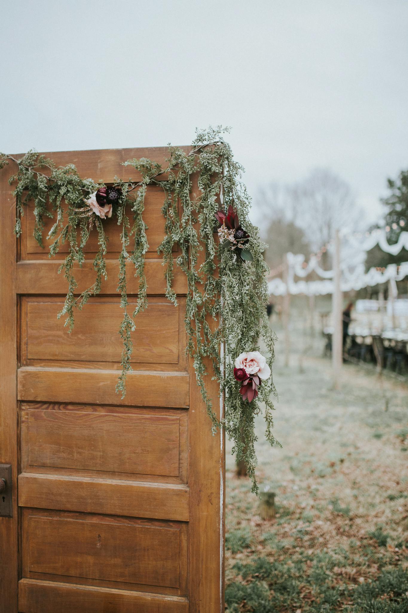 About A rustic wooden door decorated with flowers and greenery serves as an outdoor wedding entrance.